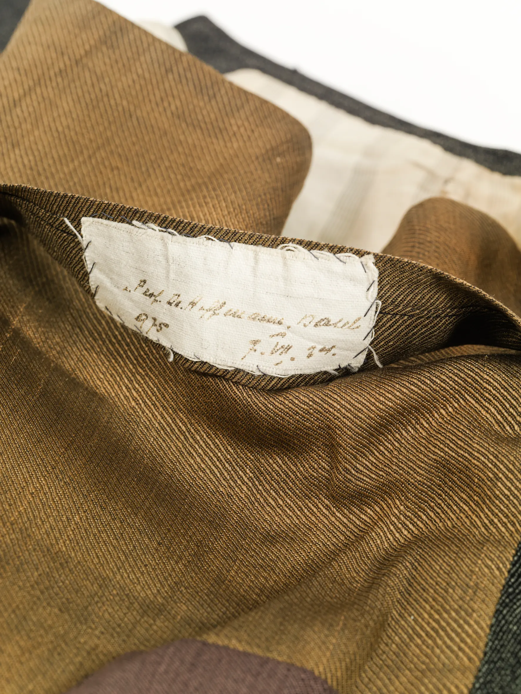 Bei einem gefalteten, braun-beigen Kleidungsstück ist ein Teil eines Saumes hochgeklappt. Dort ist mit groben Stichen eine weisse Etikette angehängt, auf der in schwarzer Schrift steht «Prof. Dr. Hoffmann, Basel». Darunter steht die Ziffer 975 und daneben das Datum 7. VII. 24.