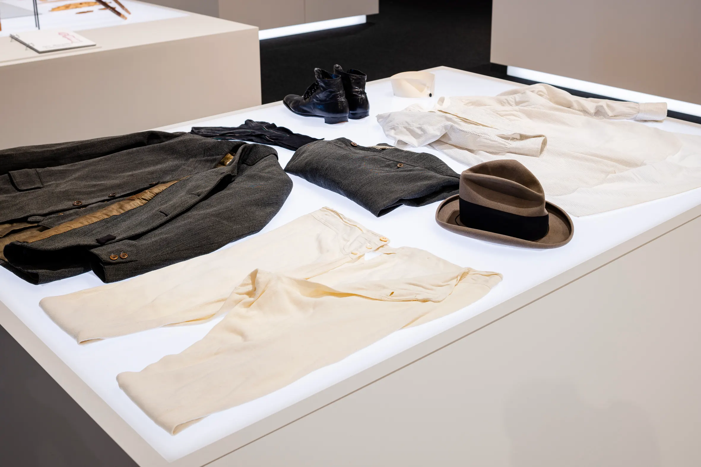 Auf einem Podest liegen verschiedene Kleidungsstücke. Links ein grauer Kittel mit glänzendem braunem Futter, rechts daneben eine weisse Unterhose mit langen Beinen und drei Knöpfen am Schnitt. In der Mitte des Podests liegen in einer Reihe von links nach rechts ein paar schwarze Socken, eine graue zusammengefaltete Hose und ein brauner Hut mit Krempe und schwarzem Band. Im oberen Teil des Podest steht ein paar glänzende schwarze Schuhe, daneben ein weisser Hemdkragen und rechts daneben liegt ein weisses Hemd mit schwarzen Nadelstreifen.
